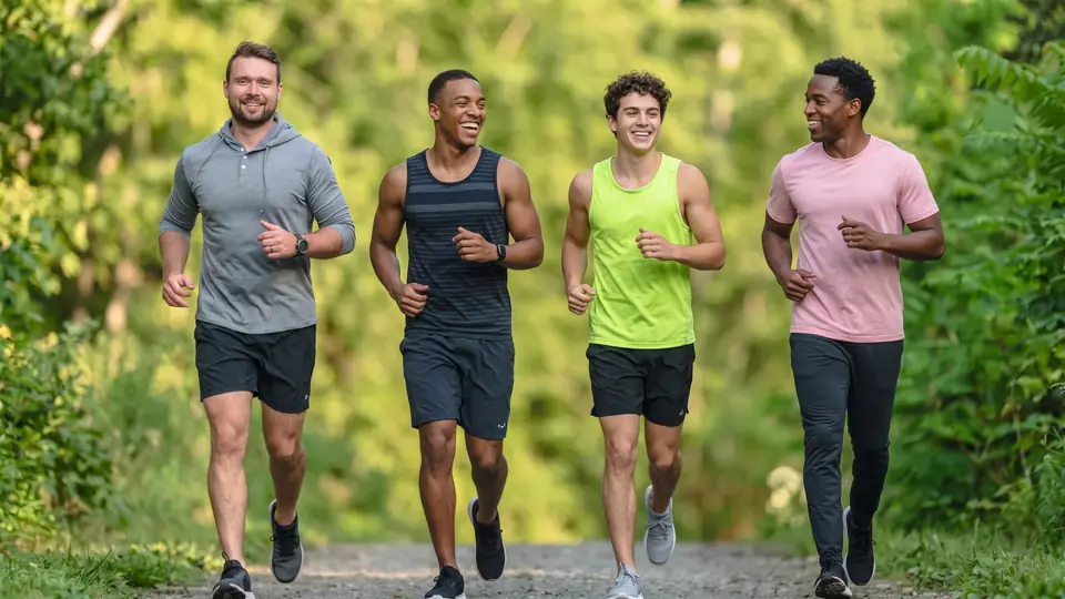 Four smiling young men jog together on a gravel path lined with green trees.