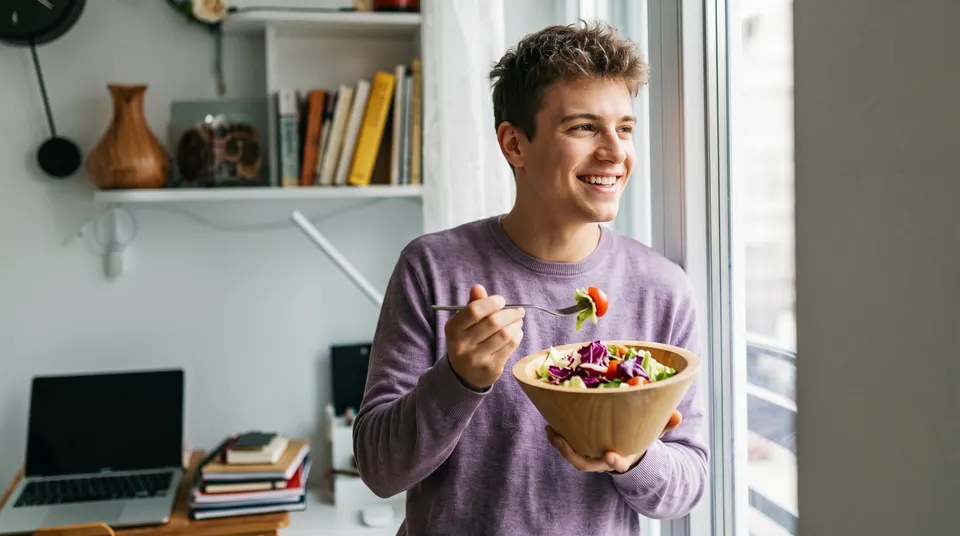 A smiling young man eats a colorful salad by an apartment window.